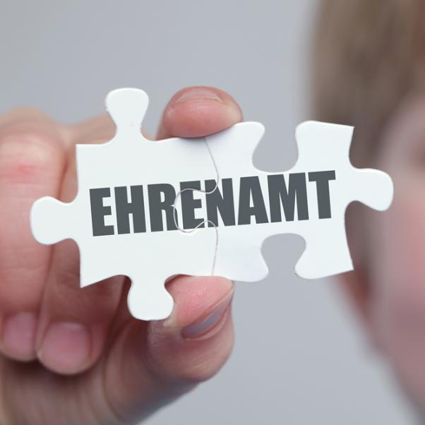 Das Foto zeigt das Gesicht eines Jungen, der zwei Puzzleteile in die Kamera hält. Auf denen steht 'Ehrenamt' geschrieben.