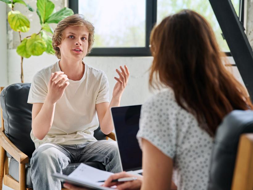 Das Bild zeigt einen sprechenden jungen Mann, der in einer Beratungssituation eine Frau gegenüber sitzt. Sie hört ihm zu.