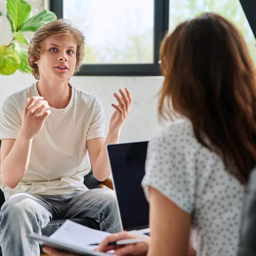 Das Bild zeigt einen sprechenden jungen Mann, der in einer Beratungssituation eine Frau gegenüber sitzt. Sie hört ihm zu.