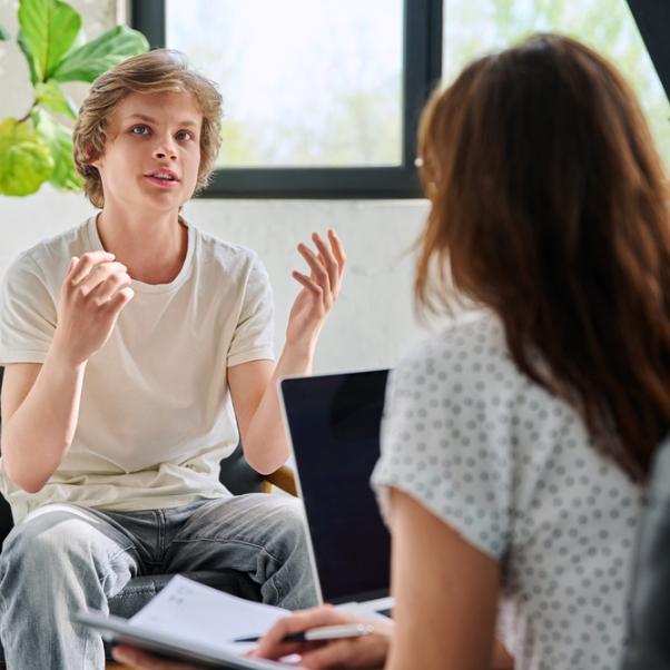 Das Bild zeigt einen sprechenden jungen Mann, der in einer Beratungssituation eine Frau gegenüber sitzt. Sie hört ihm zu.